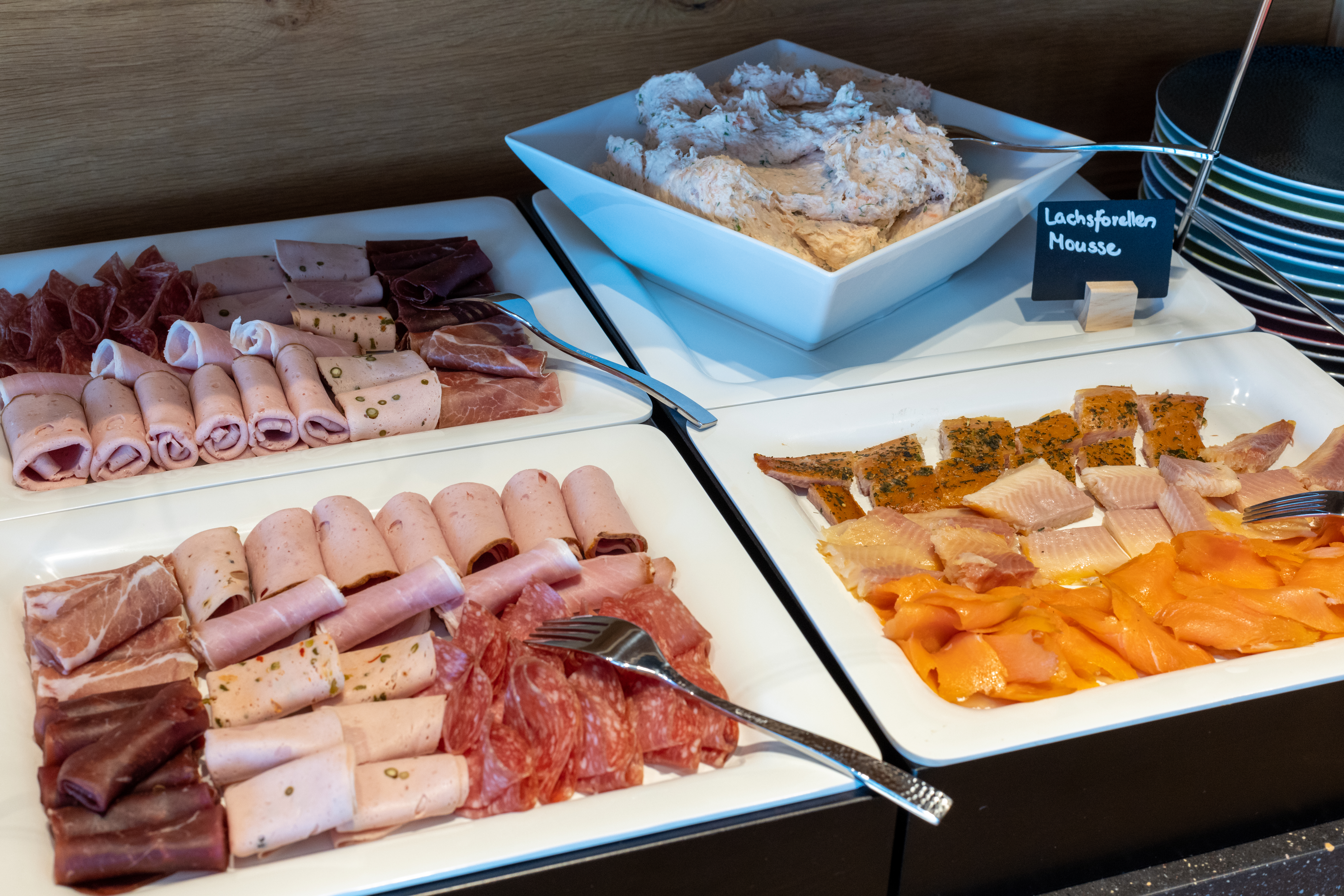 Auf einem Buffet stehen weisse Platten mit Speisen: Wurst- und Fleischplatten, Lachsforellenmousse sowie geräucherte Lachsforelle und Forelle. Ein Schild "Lachsforellen Mousse" steht daneben. Teller sind gestapelt.